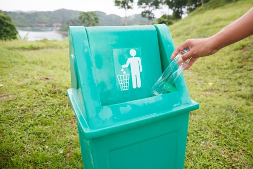 A person is seen disposing of a clear plastic bottle into a bright turquoise recycling bin situated outdoors on a grassy area. The bin features a white icon of a person throwing waste into a bin and is positioned in an open space with a scenic background that includes trees, a body of water, and distant hills, suggesting a park or lakeside setting. The scene is illuminated by natural daylight, highlighting the clean, smooth surface of the bin and the textured grass underneath. The individual’s hand, visible on the right side, is placing the bottle into the designated opening of the bin. This image emphasizes environmentally responsible waste disposal practices, aligning with rubbish removal services offered by Garden Clearance Stoke Newington in the local Stoke Newington area, serving nearby communities within N16 postcode district.