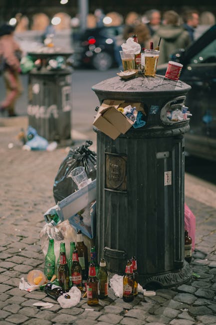 A crushed aluminum energy drink can with a predominantly blue and black design lies on a dark, textured asphalt surface. Next to it is a small, folded piece of white paper or cardboard with a maze-lik