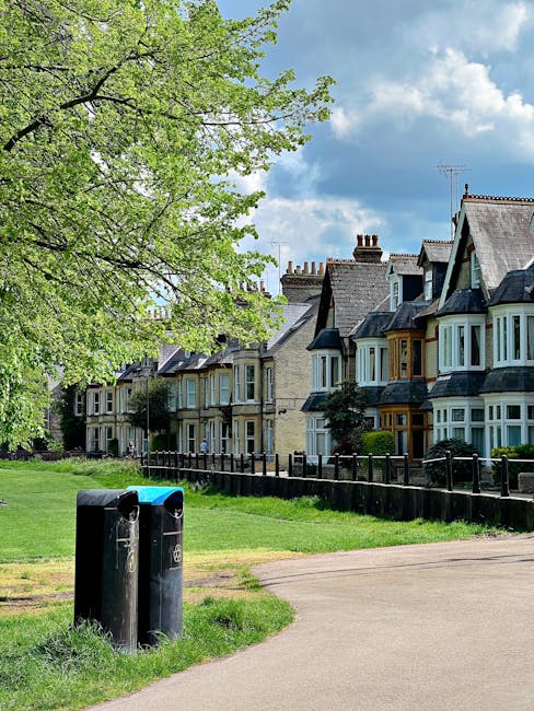 In the foreground, there are two black public litter bins placed along a paved pathway, with a small patch of grass nearby. Behind the bins, a well-maintained lawn extends towards a row of Victorian-s