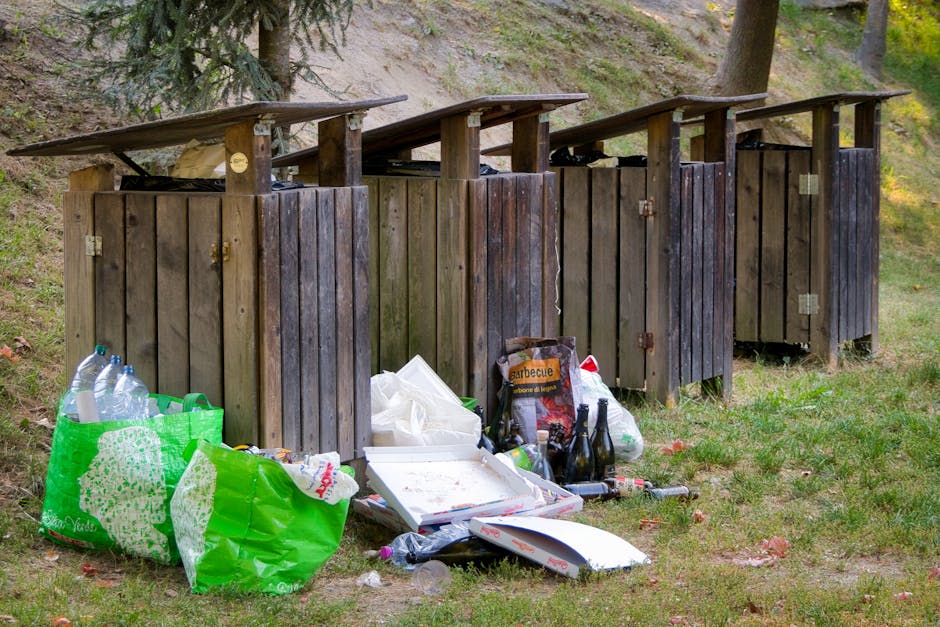 In the foreground, there are two black public litter bins placed along a paved pathway, with a small patch of grass nearby. Behind the bins, a well-maintained lawn extends towards a row of Victorian-s