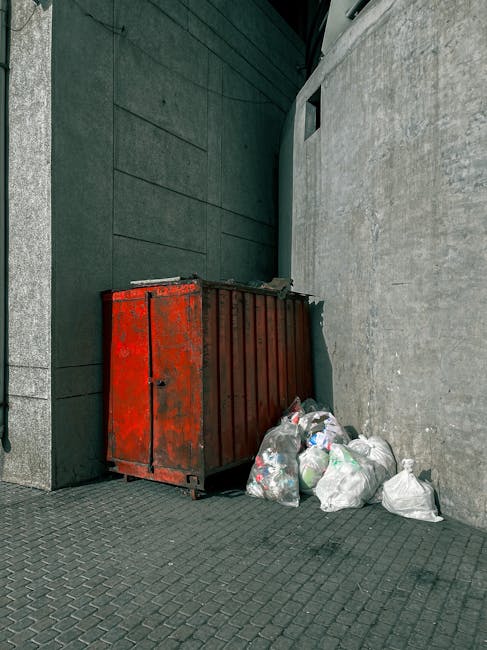 A cluttered urban alleyway scene shows a large, black garbage bag in the foreground, situated on a concrete pavement stained with dark patches. Behind the bag, a metal trolley or cart is partially vis