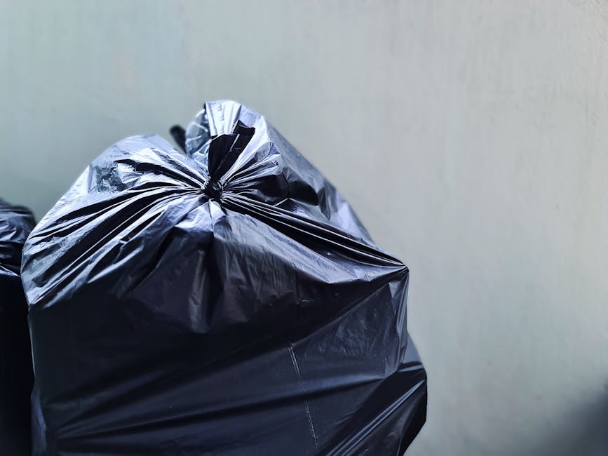 Close-up view of tightly knotted black plastic rubbish bags, made of shiny, smooth material, stacked against a plain light-colored background. The bags appear full and are positioned in the foreground